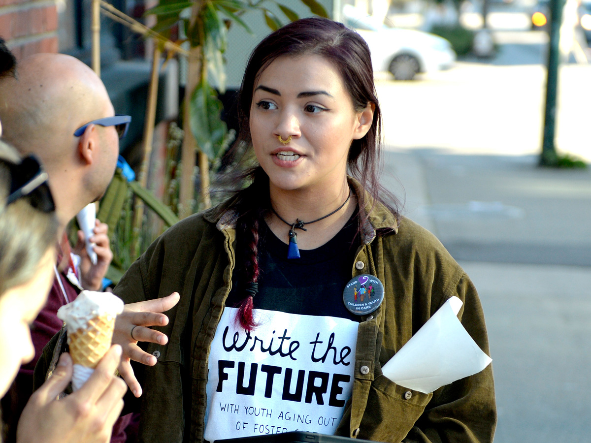 A young woman with dark braided hair wearing a dark shirt with the text "Write the Future with Youth aging out of foster care" speaking to people on a sidewalk